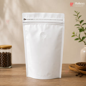 A blank white stand-up resealable pouch with a zipper closure and a small circular vent hole, standing on a wooden surface. The background is a warm beige wall with soft shadows, and decorative props including a jar of coffee beans and a small plant are visible on both sides.