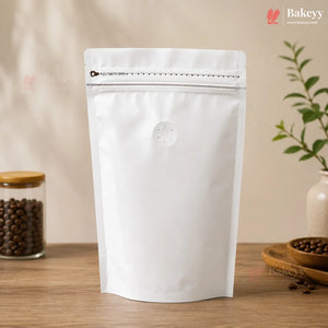 A blank white stand-up resealable pouch with a zipper closure and a small circular vent hole, standing on a wooden surface. The background is a warm beige wall with soft shadows, and decorative props including a jar of coffee beans and a small plant are visible on both sides.
