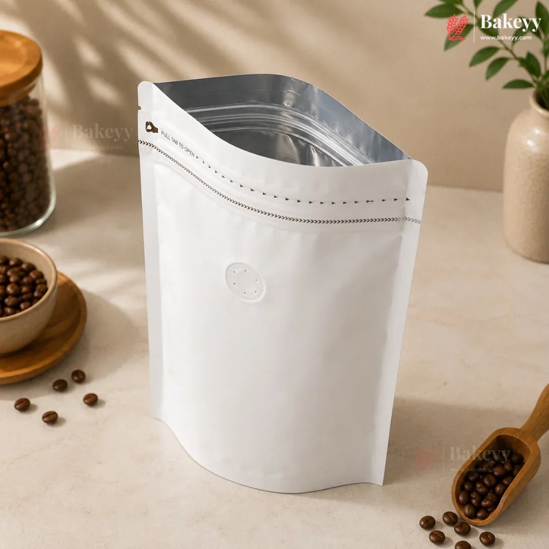 A white stand-up resealable coffee bag with a silver zipper, styled on a beige tabletop with coffee beans, a wooden scoop, and a small plant in the background.