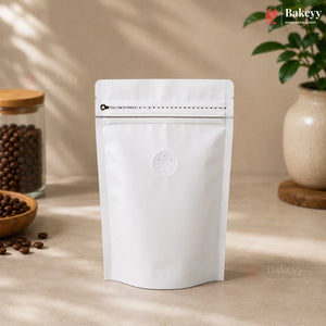 A blank white stand-up resealable pouch with a zipper closure and a small circular die-cut window near the bottom center, standing on a beige tabletop. Coffee beans in jars and a potted plant are softly blurred in the background with warm natural shadows.