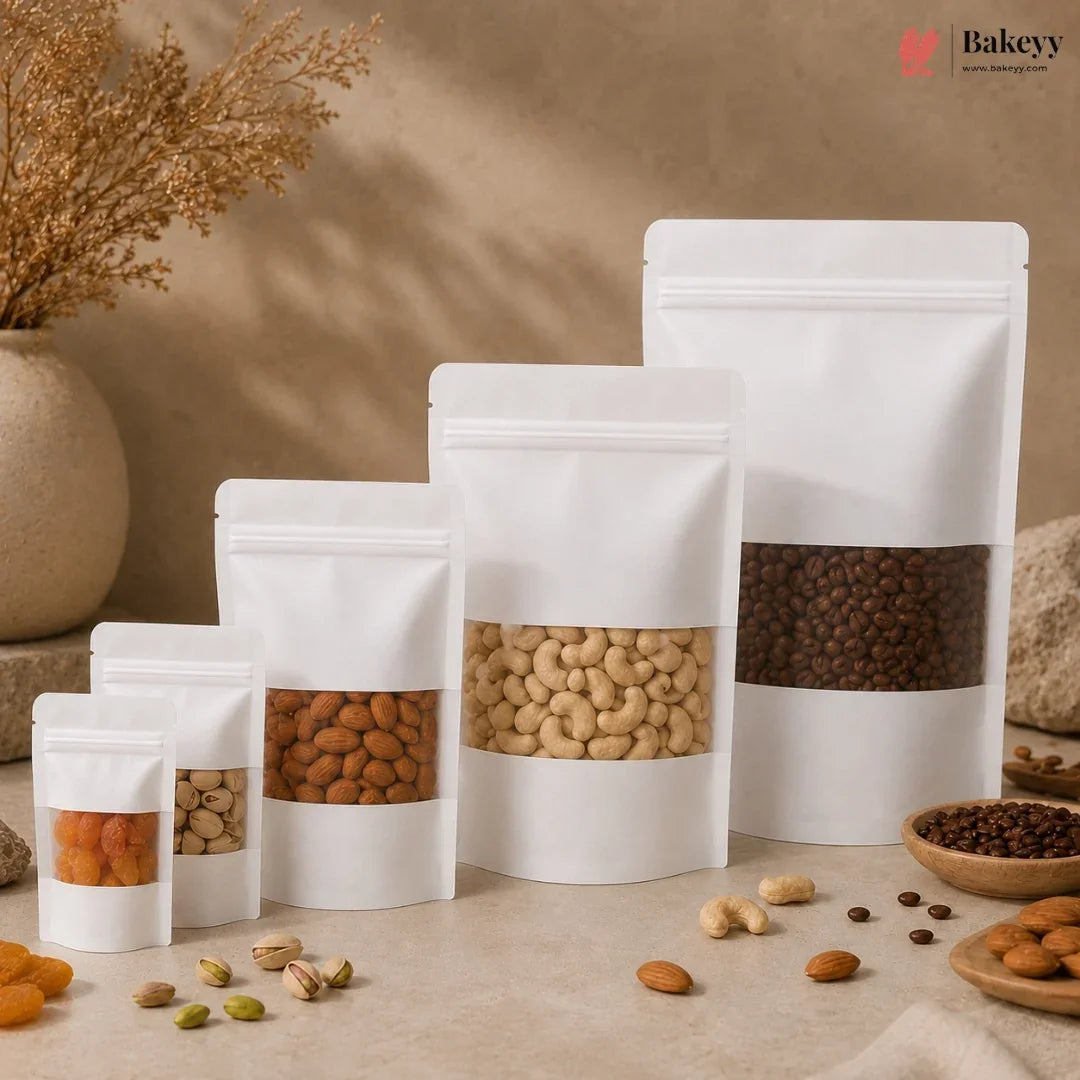 Five white stand-up resealable pouches of different sizes with clear windows displaying various nuts and dried fruits, arranged on a beige surface with decorative dried flowers and bowls of snacks in the background.