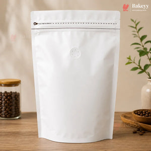 A white stand-up resealable pouch with a zipper seal and a small circular vent hole, displayed on a wooden surface with coffee beans and a plant in the background. The Bakeyy logo is visible in the top corners.