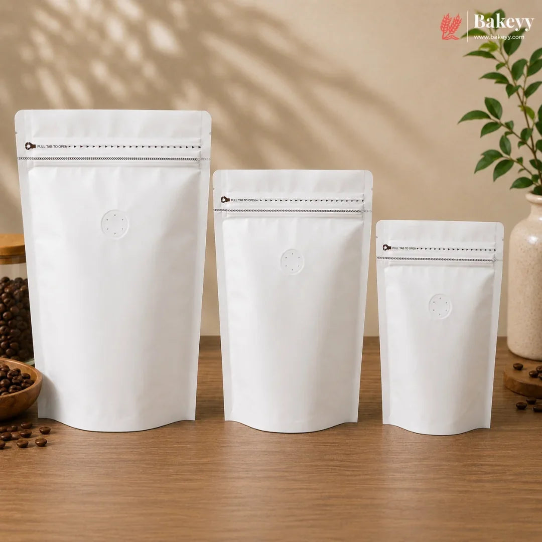 Three white stand-up pouches in different sizes arranged on a wooden table, with coffee beans and a plant in the background. The Bakevy logo appears in the top right corner.