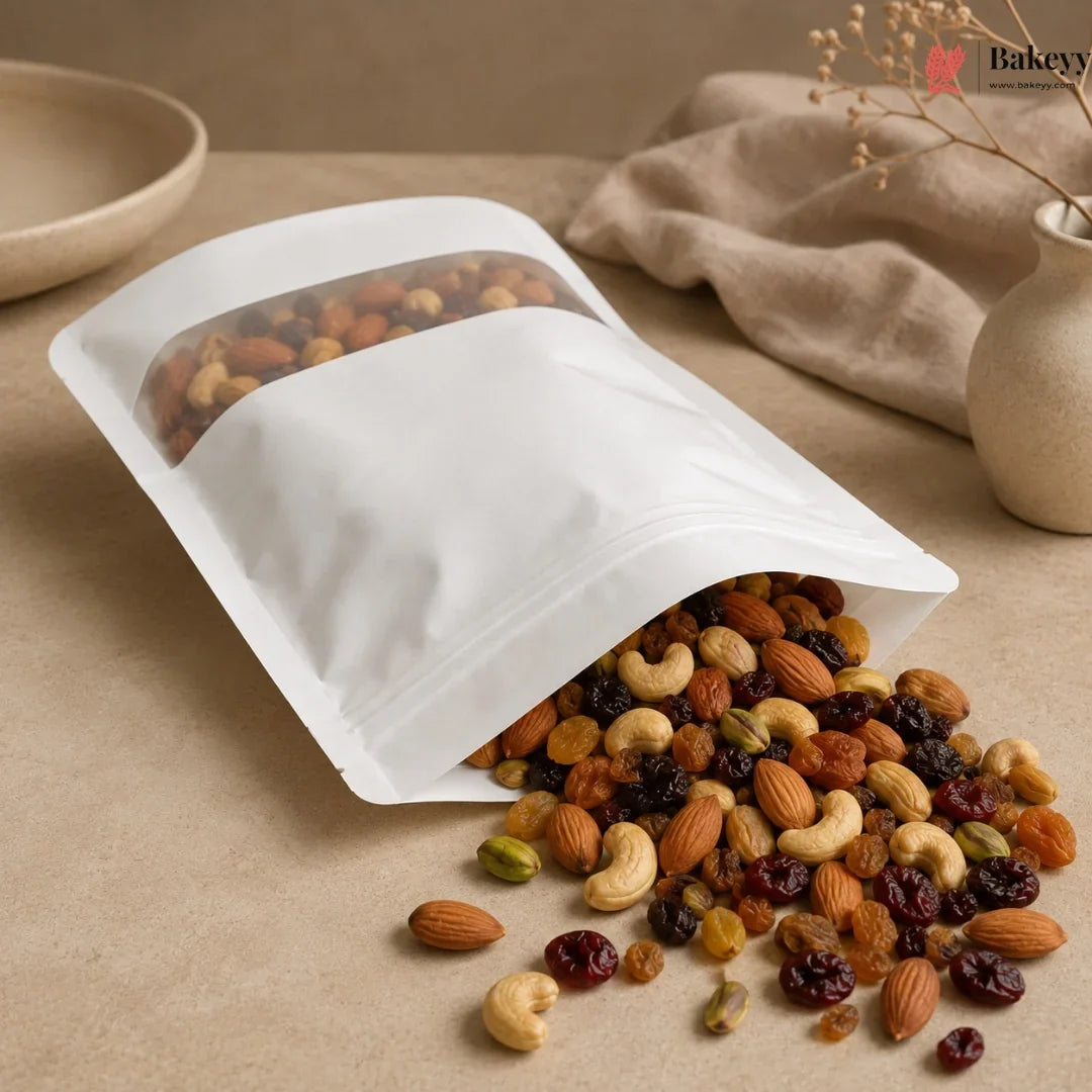 A white stand-up resealable pouch with a clear window, shown on a beige tabletop with mixed nuts and dried fruit spilling out. A beige cloth and ceramic vase are in the background, and a Bakeey logo appears in the top right corner.