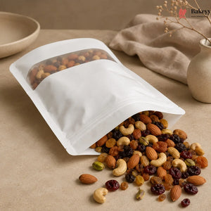 A white stand-up resealable pouch with a clear window, shown on a beige tabletop with mixed nuts and dried fruit spilling out. A beige cloth and ceramic vase are in the background, and a Bakeey logo appears in the top right corner.