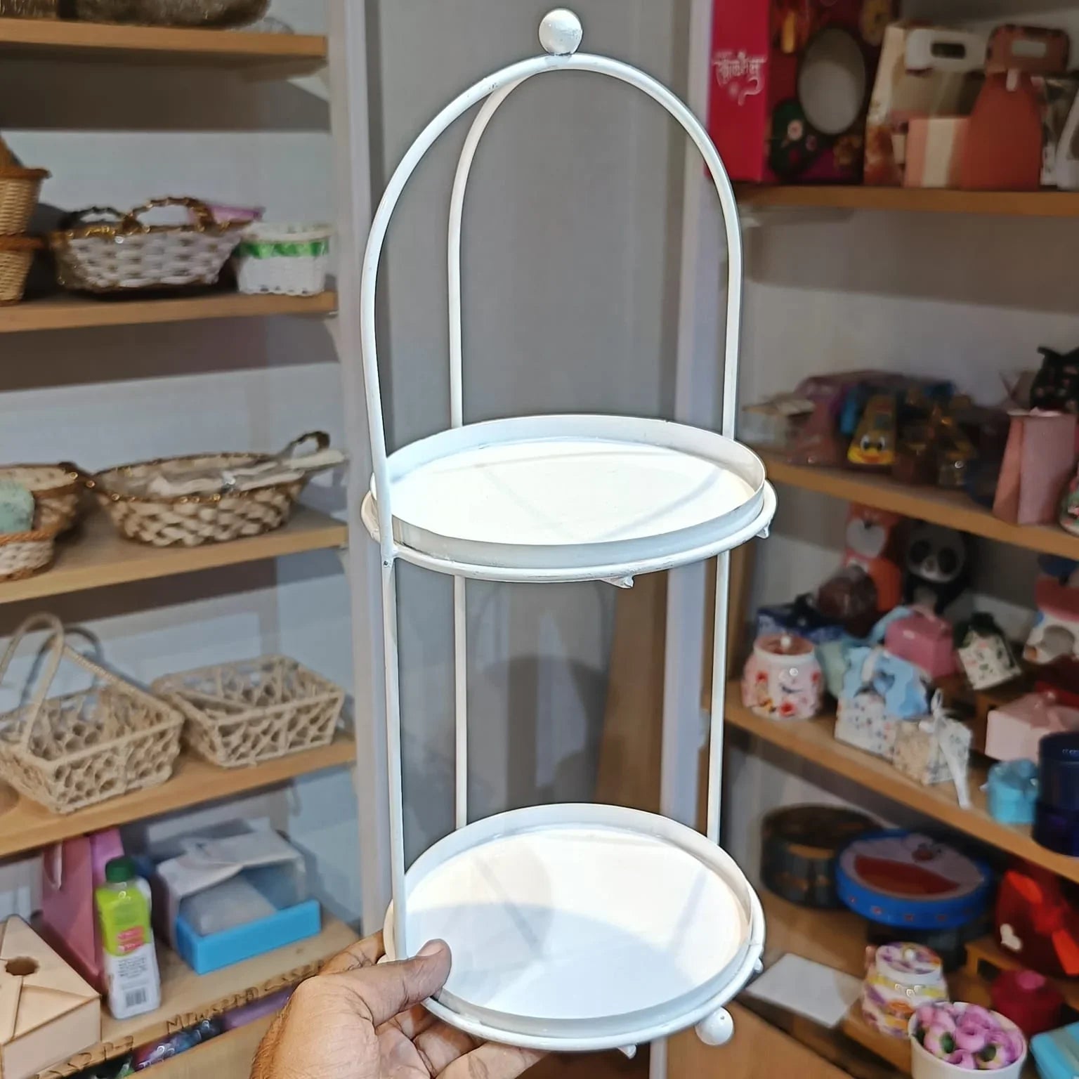 Two-tiered white metal stand held by a hand with shelves in the background
