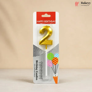 A packaged gold birthday candle shaped like the number 2, shown on its retail card with 'HAPPY BIRTHDAY' text and balloon graphics. The package is photographed against a soft beige background.