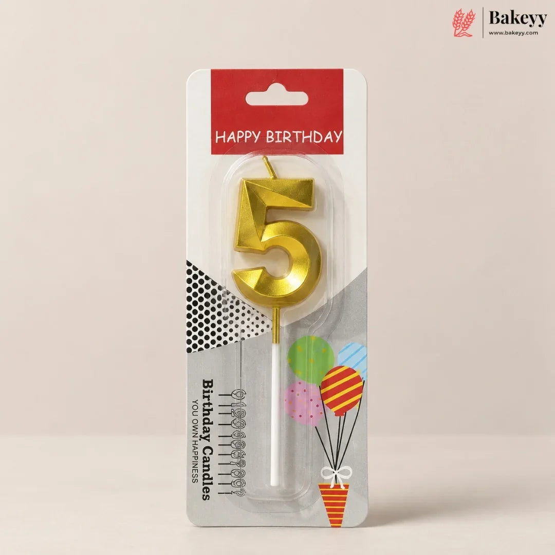 A gold metal candle shaped like the number 5 for birthdays, packaged in a clear blister pack on a white and red card that reads 'HAPPY BIRTHDAY' and shows birthday-themed graphics. The product is photographed against a plain beige background.
