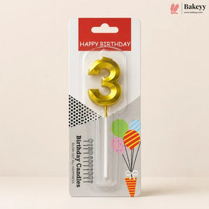 A gold metallic number 3 birthday candle in its original retail packaging, featuring a red 'HAPPY BIRTHDAY' header and festive balloon graphics on a light beige background.