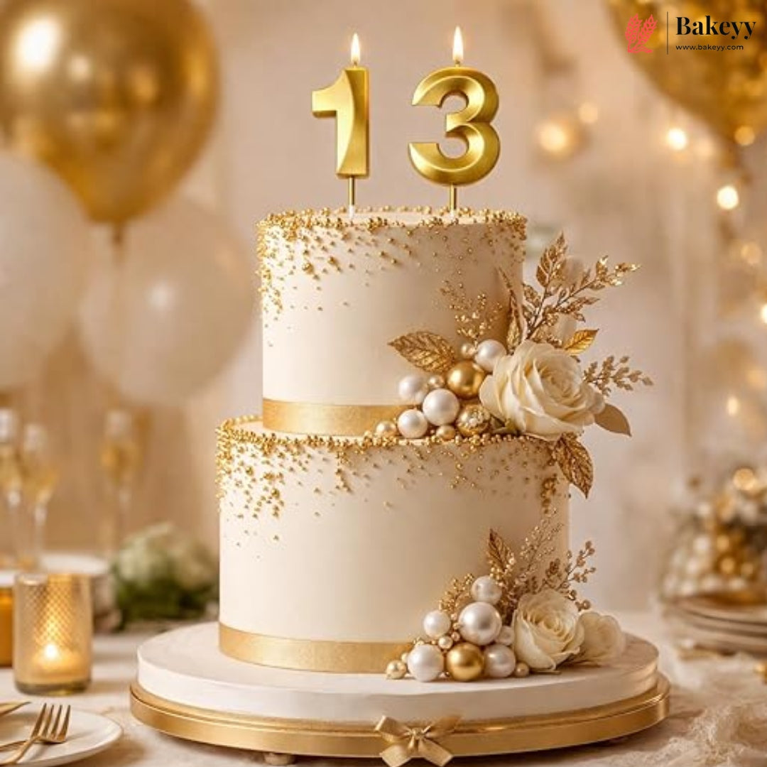 A two-tier birthday cake decorated in elegant white and gold, topped with gold number 13 candles, pearls, and white roses, set on a decorated party table.