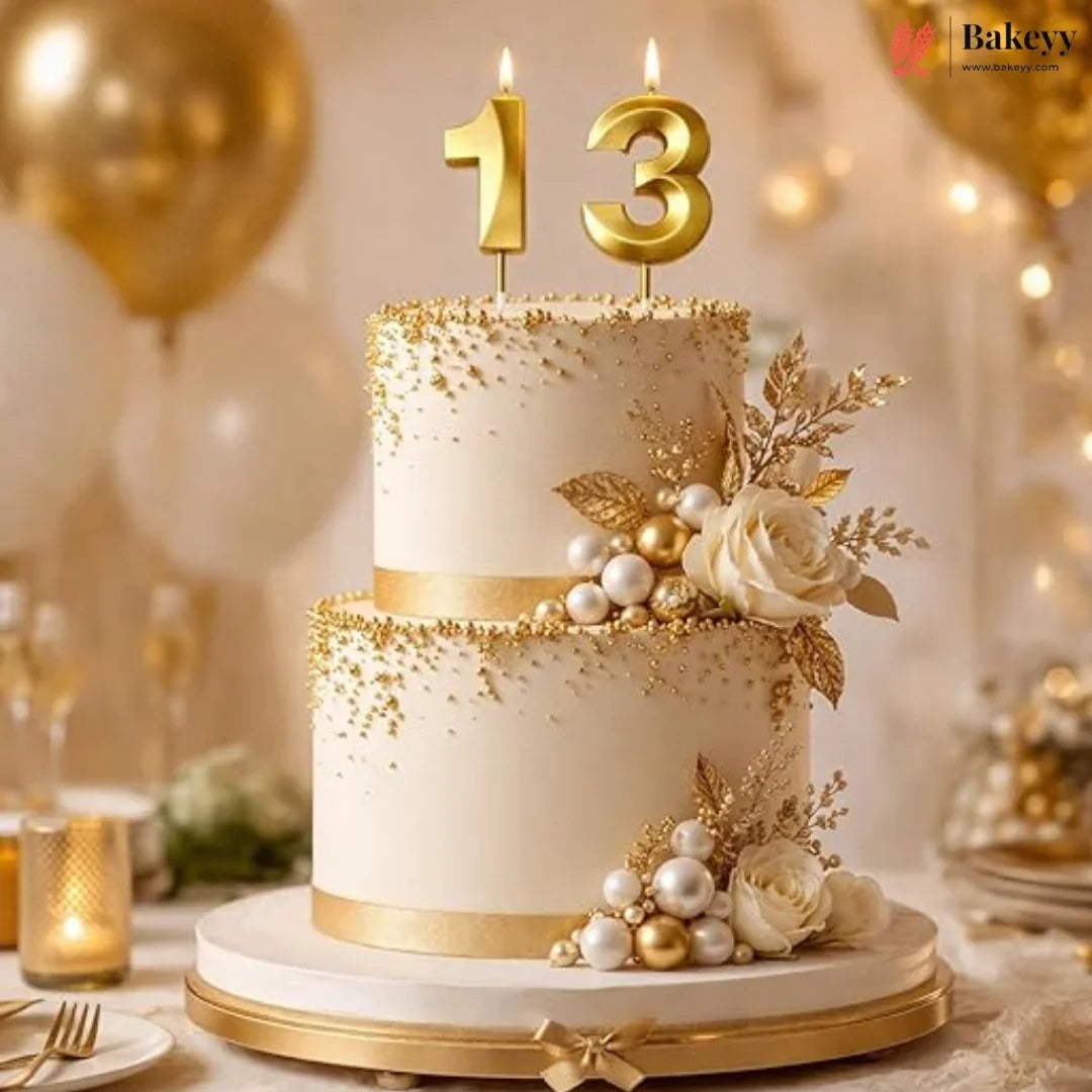A two-tier birthday cake decorated in elegant white and gold, topped with gold number 13 candles, pearls, and white roses, set on a decorated party table.
