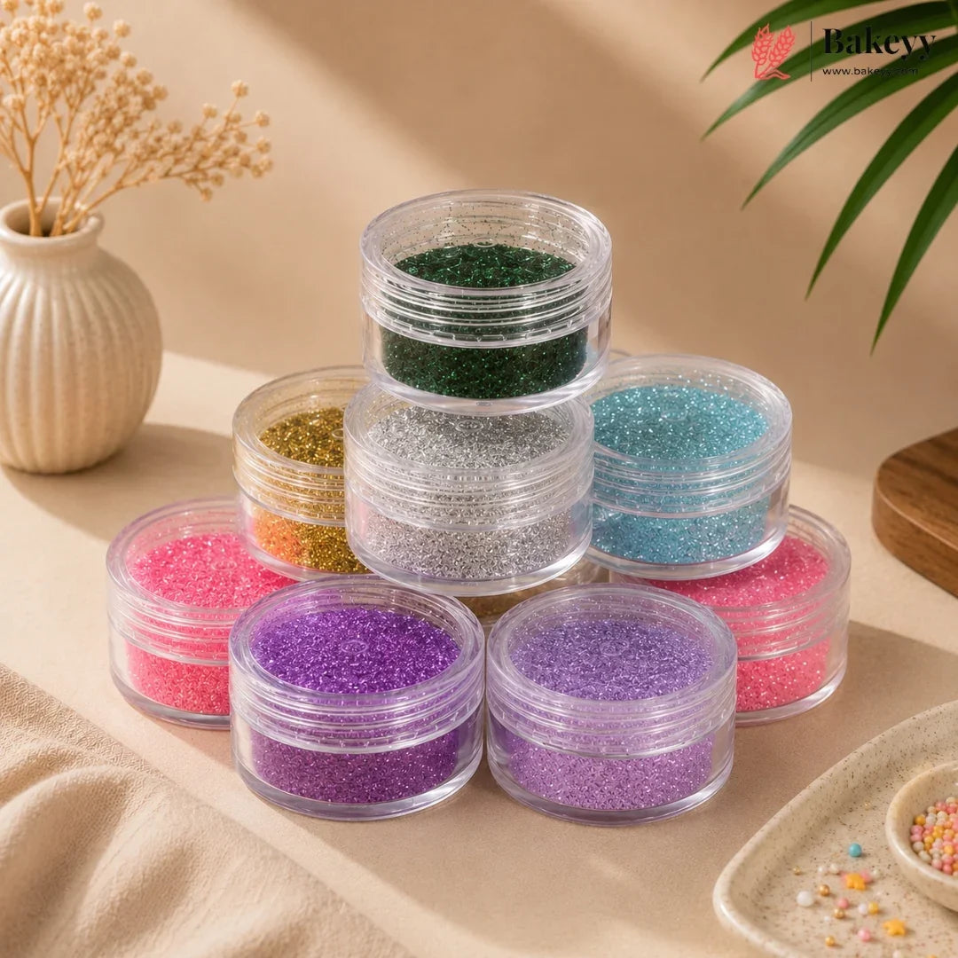 A pyramid arrangement of small clear plastic jars filled with colorful glitter, displayed on a beige fabric surface with decorative props including a vase with dried flowers, a green leaf, and a small dish of sprinkles.