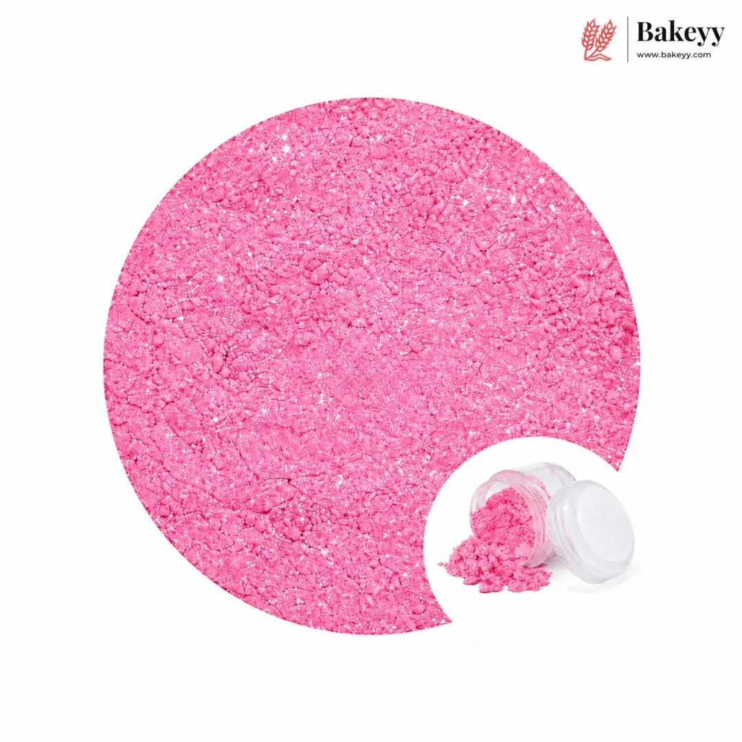 Bright pink edible glitter or sugar crystals shown in a large circular close-up on a white background, with a small inset of an open clear jar containing more of the pink powder. The Bakeyy logo appears in the top right corner.
