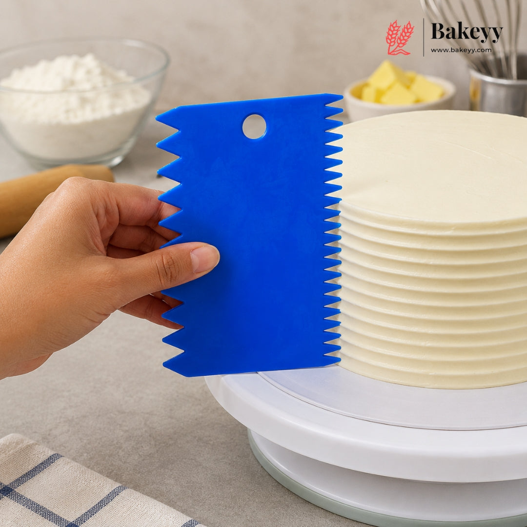 A hand holds a bright blue serrated cake scraper against a white frosted cake on a rotating cake stand, with bowls of flour and butter blurred in the background. The Bakeyy logo appears in the top right.