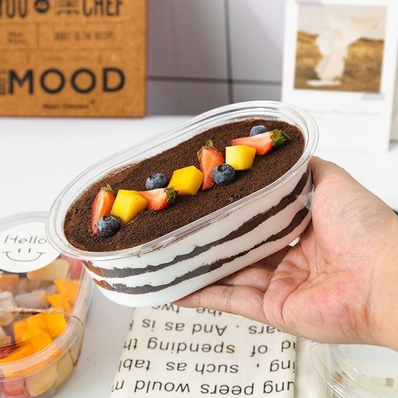 Hand holding a dessert container with fruit toppings on a table with a magazine underneath.