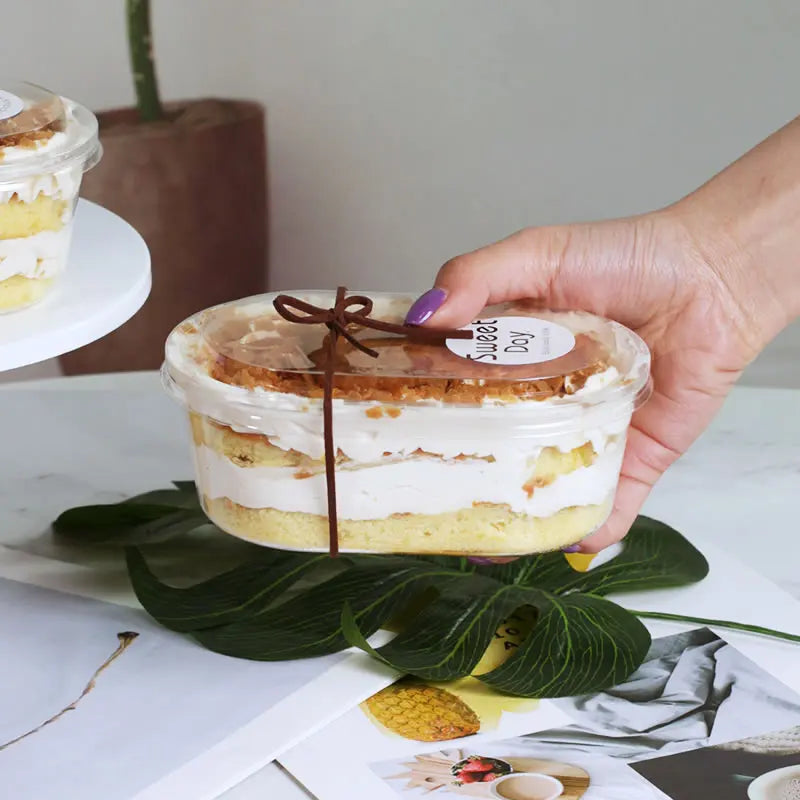 Hand holding a layered dessert in a transparent container with a decorative bow on a light surface.