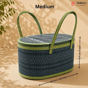 A medium-sized woven basket with a dark blue-gray body, green trim, and two green braided handles, shown with dimension labels on a beige background with leaf shadows. The Bakeyy brand logo is visible in the top right.