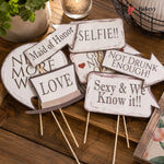 Set of humorous wooden signs on sticks with various sayings on a wooden surface.