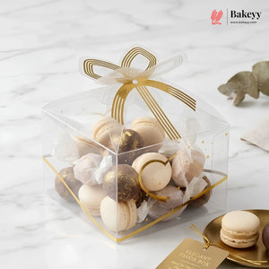 Clear favor box with macarons and gold ribbon on a marble surface, featuring the Bakeyy brand.