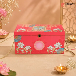 A rectangular red decorative jewelry box with floral and geometric patterns, featuring a gold clasp and a pink oval detail on the front, styled on a beige surface with flowers and candles.