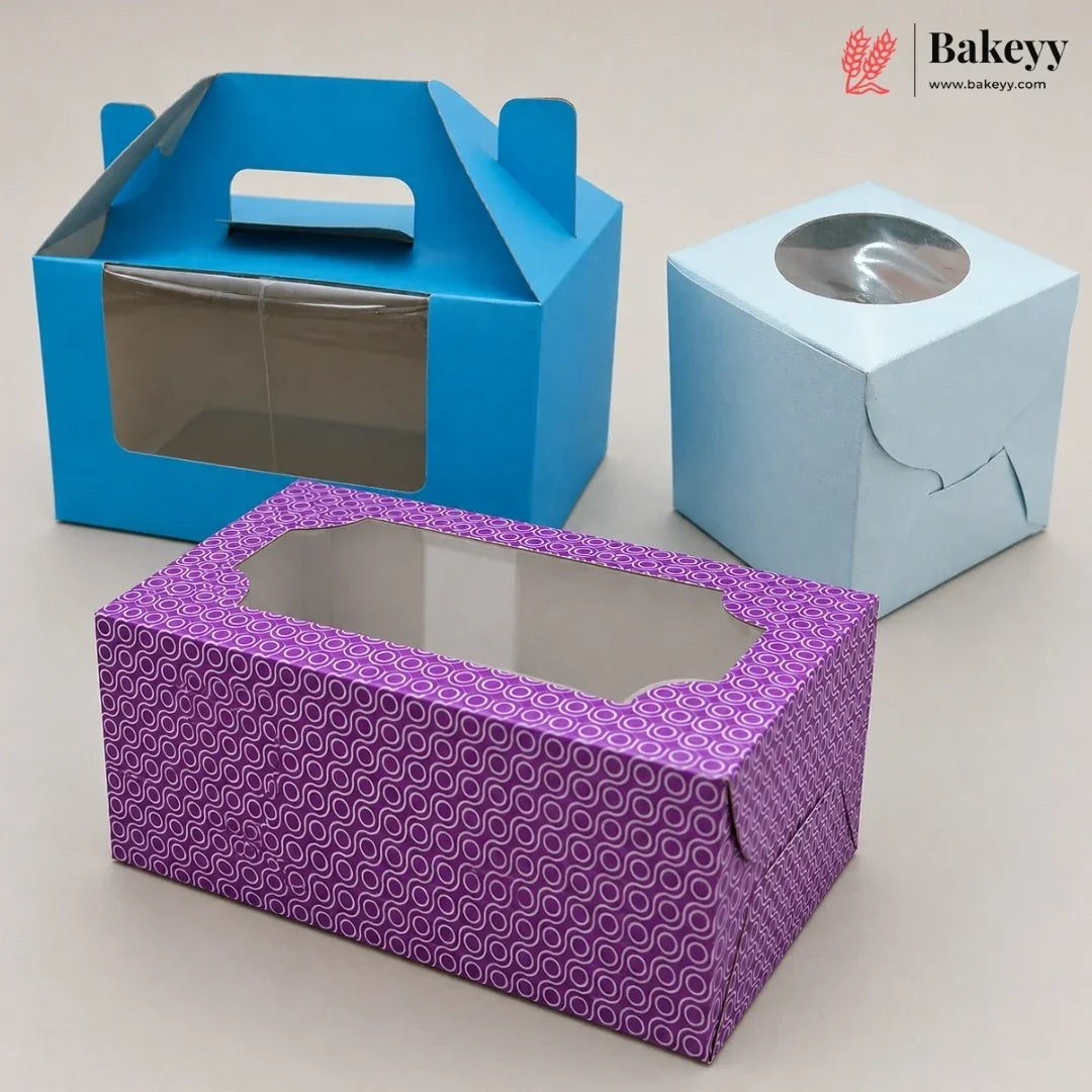 Three paper bakery boxes on a light beige background: a blue handled box with a clear window, a light blue cube with a round clear window, and a purple patterned rectangular box with an octagonal clear window. A Bakeyy logo appears in the top right corner.
