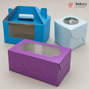 Three paper bakery boxes on a light beige background: a blue handled box with a clear window, a light blue cube with a round clear window, and a purple patterned rectangular box with an octagonal clear window. A Bakeyy logo appears in the top right corner.
