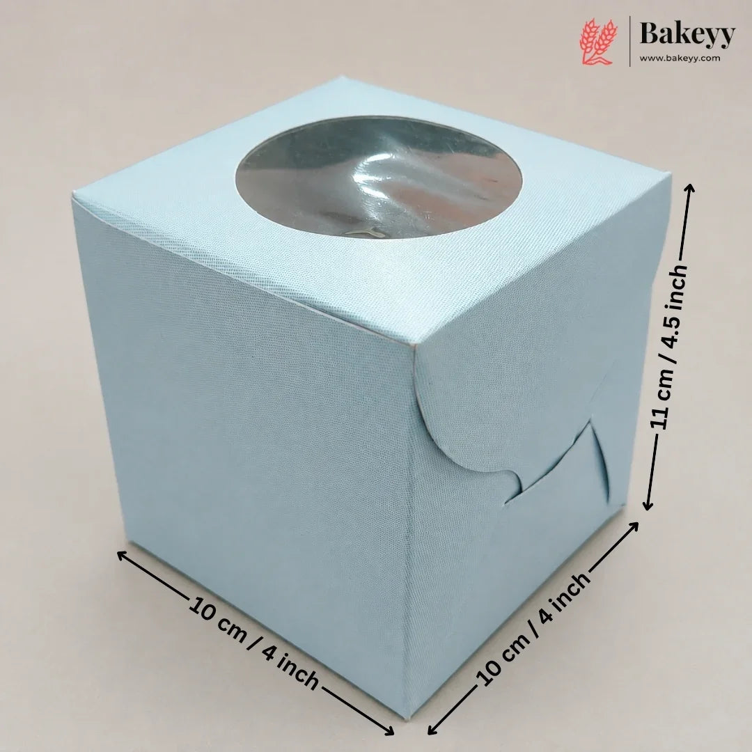 A light blue square paper gift box with a circular clear window on top, shown with dimension arrows and labels indicating 10 cm by 10 cm by 11 cm measurements. The Bakeyy logo appears in the top right corner.