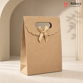 A kraft paper gift bag with a scalloped flap, a cutout handle, and a tied ribbon bow, photographed in a warm beige studio setting with soft shadows. The Bakeyy logo appears in the top right corner.
