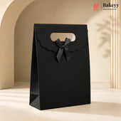 A black paper gift bag with scalloped handle edges and a matching black ribbon bow, standing on a beige surface in a softly lit studio setting. The Bakeyy logo appears in the top right corner.