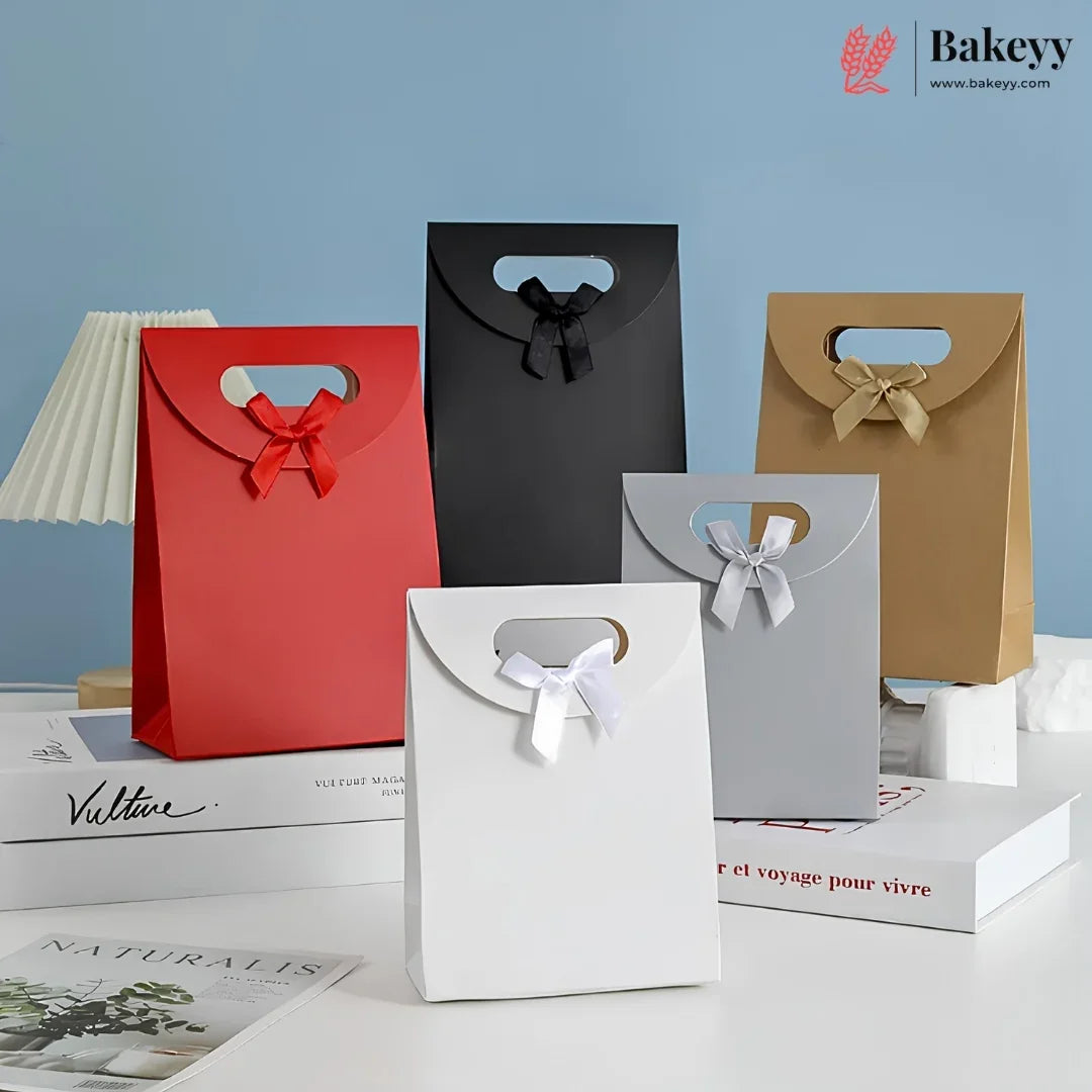 Five paper gift bags in red, black, brown, white, and gray, each with a handle cutout and a ribbon bow at the top, arranged on books against a light blue background. The Bakeyy logo appears in the top right corner.