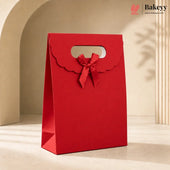 A red paper gift bag with a scalloped handle and a matching red ribbon bow, standing on a beige surface in a softly lit studio setting. The Bakeyy logo appears in the top right corner.