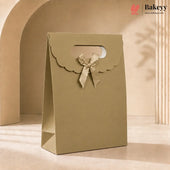 A kraft paper bakery gift bag in a warm beige-tan color, featuring a scalloped handle cutout and a matching satin ribbon bow. The bag is photographed in a warm, minimalist studio setting with soft shadows.