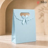 A light blue bakery gift bag with a scalloped handle and a matching ribbon bow, standing on a beige surface against a warm beige architectural background. The Bakeyy logo is visible in the top right corner.