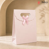 A light pink paper gift bag with a scalloped flap handle, a matching pink ribbon bow, and a cutout window near the top, photographed in a warm beige studio setting with soft shadows. The Bakeyy logo appears in the top right corner.