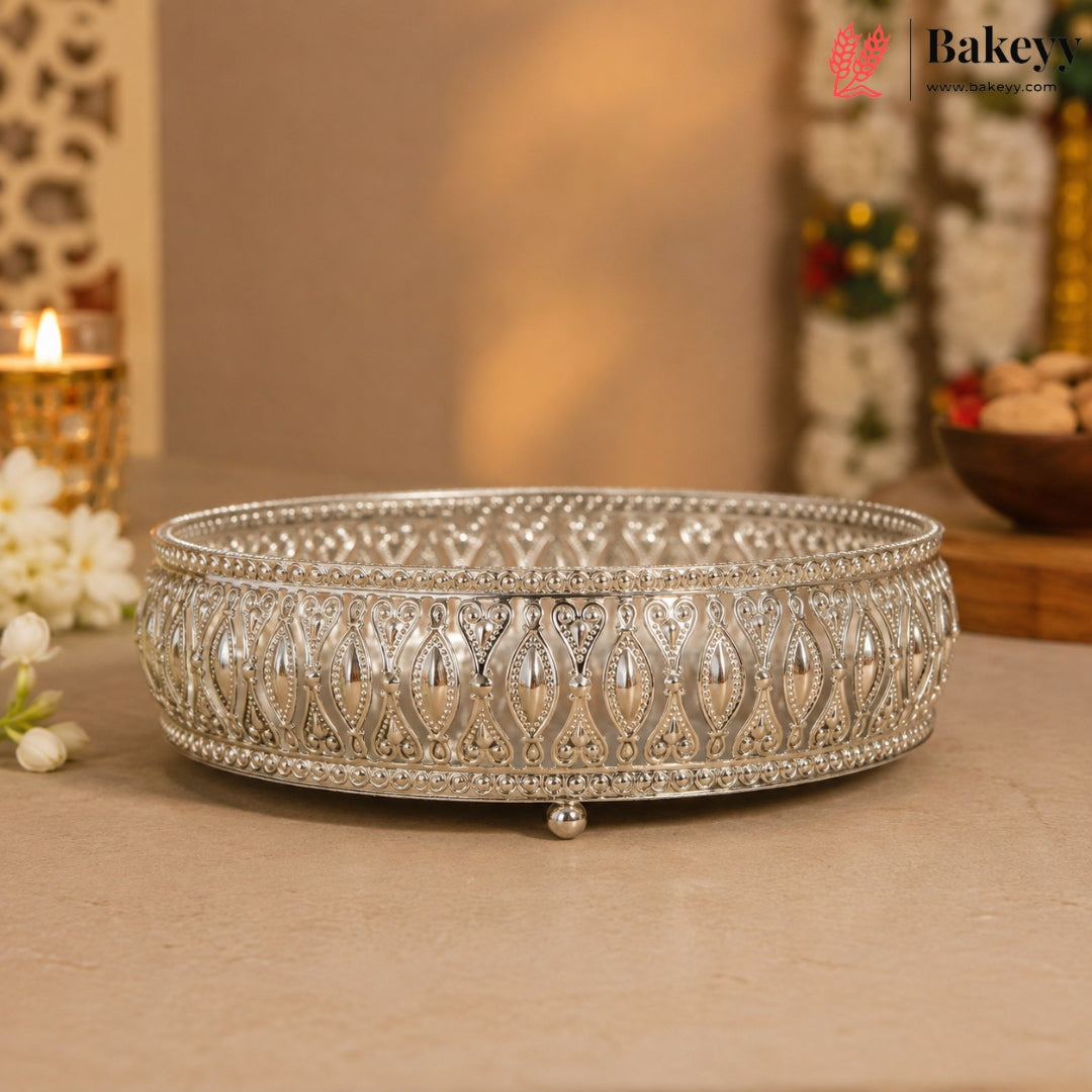 An ornate silver-colored metal tray with intricate filigree patterns and small feet, displayed on a beige surface with a warm, softly blurred background.