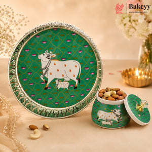 A decorative green and silver plate with a cow and calf illustration, shown beside a matching green lidded container filled with almonds on a warm beige surface. The Bakeyy brand logo appears in the top right corner.