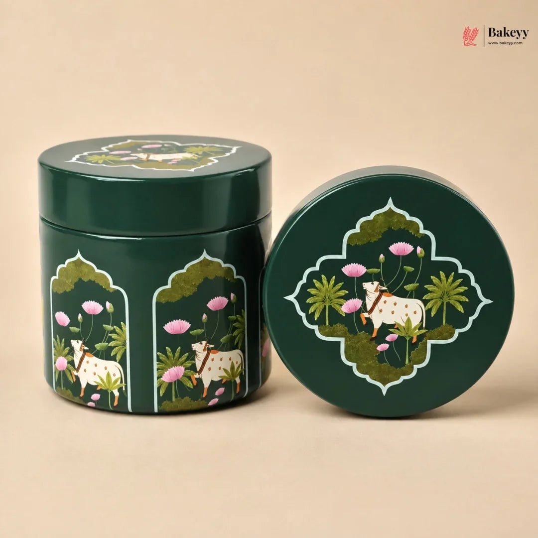 A dark green round metal tin with a matching lid, decorated with an intricate hand-painted style illustration of a white goat, pink lotus flowers, and green foliage. The tin is shown standing next to its lid on a solid beige background.
