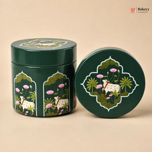 A dark green round metal tin with a matching lid, decorated with an intricate hand-painted style illustration of a white goat, pink lotus flowers, and green foliage. The tin is shown standing next to its lid on a solid beige background.