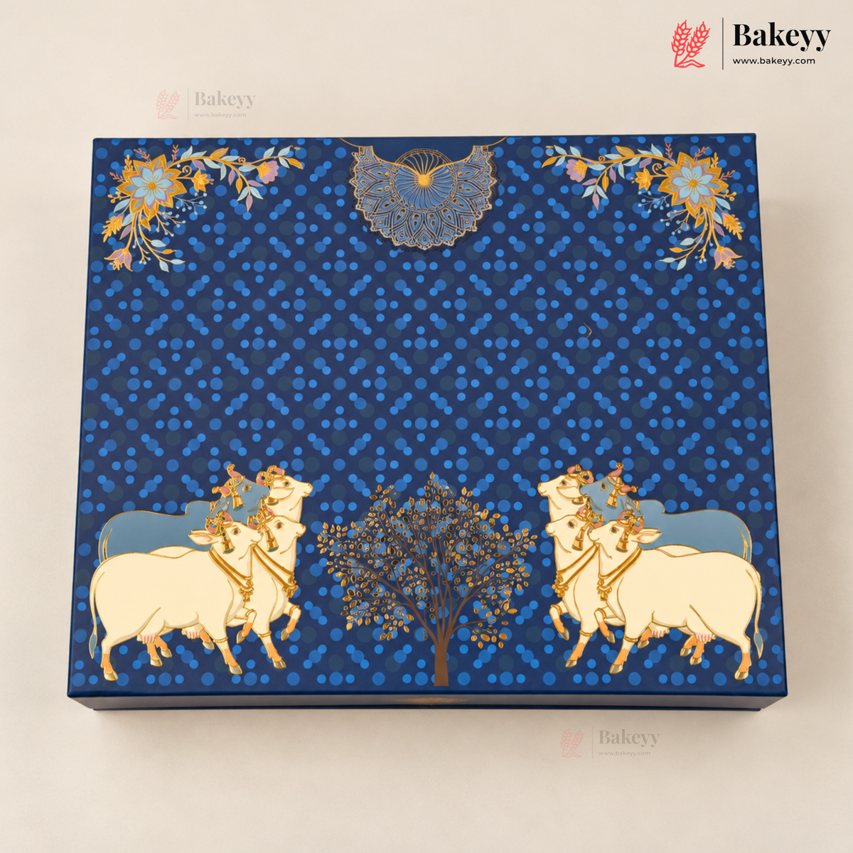 A square blue decorative box with intricate gold and white illustrations of cows, a tree, floral motifs, and a bird, photographed on a light beige background. The Bakeyy logo is visible in the top right corner.