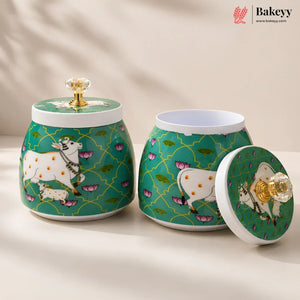 Two decorative ceramic canisters with matching lids on a warm beige surface. The canisters have a teal green background with a gold trellis pattern, pink lotus motifs, and white cows with yellow spots.