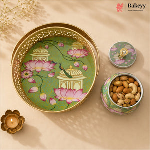 A decorative round metal tray with a gold-tone rim and handle, shown alongside its matching lid and a small open container filled with almonds and cashews. The tray features an illustrated design of pink lotus flowers, domes, and a peacock on a green background.