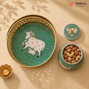 A decorative ceramic and metal tin set with a cow and calf motif on a teal background, shown open with nuts inside. The set includes a large tray, a small jar, and a lid, styled on a beige surface with a diya and leaf shadows.