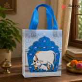 A reusable gift bag with bright blue handles sits on a wooden table in a styled indoor setting. The bag has a white base with a large blue arched illustration featuring a white cow and decorative motifs, plus a Bakeyy logo printed on the upper right side of the bag.