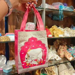 A hand holds a reusable shopping bag with pink handles and a cream-colored body featuring a large pink and white cow illustration. The background shows a retail store shelf filled with various colorful items.