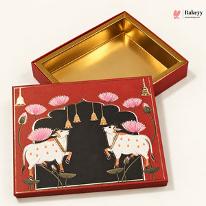 A decorative red gift box with a gold interior, shown open with the lid placed beside it. The lid features an illustrated design of two white cows with orange spots under a black arch, surrounded by pink lotus flowers and small gold bells, on a clean white background.