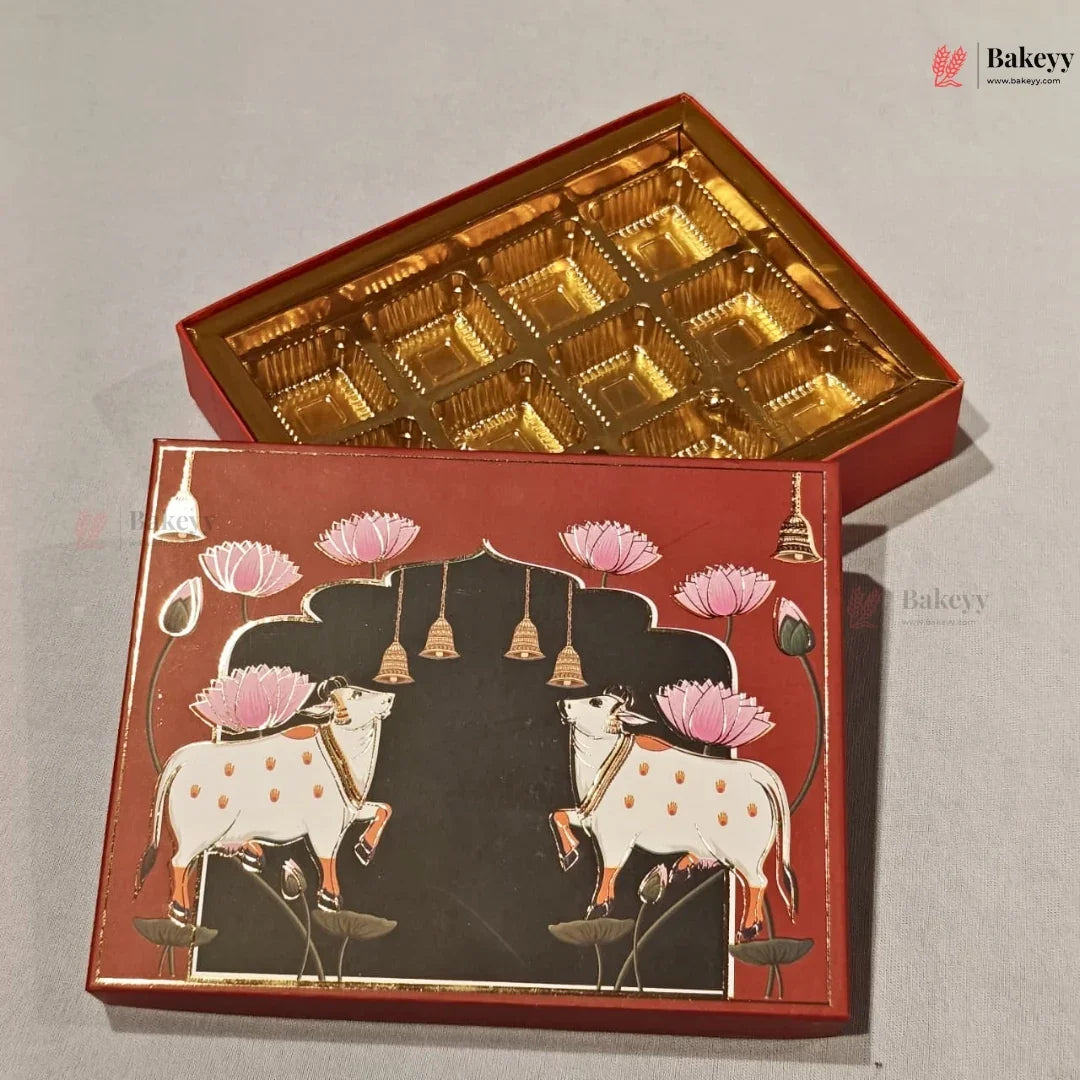 An open red and gold gift box with multiple gold tray compartments inside, shown alongside the closed front cover decorated with two white cows, pink lotus flowers, hanging bells, and a black central panel on a light gray background.