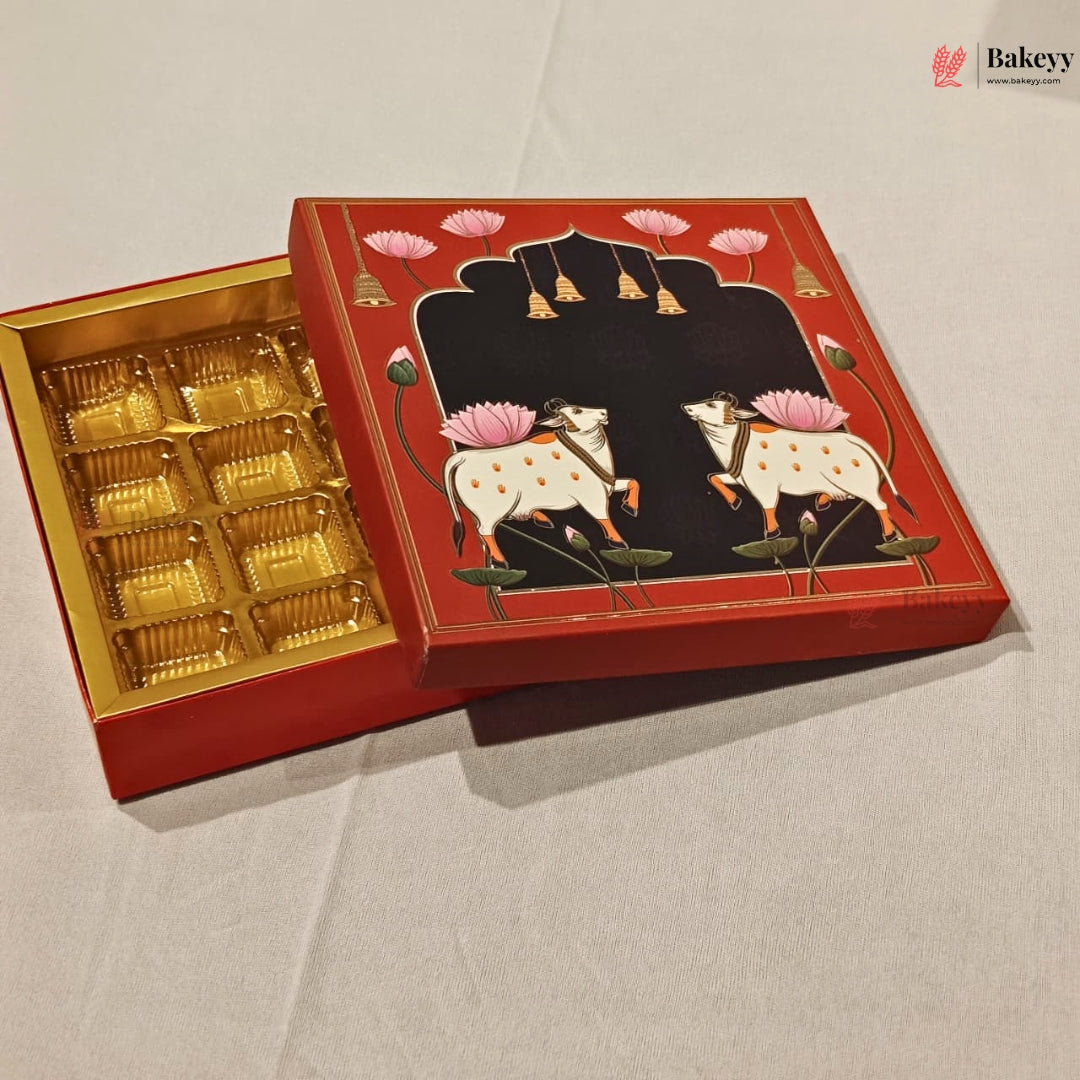 An open red gift box with a gold foil-lined interior, shown on a light fabric background. The lid features a black arched panel with illustrated white cows, pink lotus flowers, and small hanging bells.