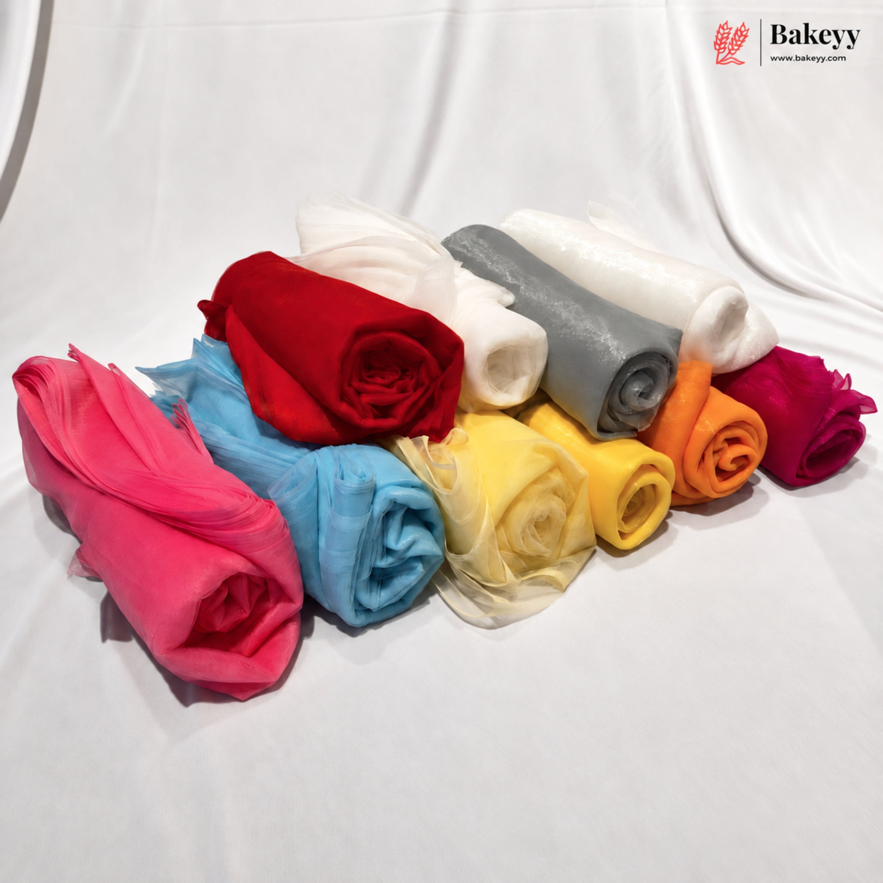 A collection of rolled sheer tulle fabric in multiple colors, including pink, blue, red, white, gray, yellow, orange, and magenta, arranged on a white draped fabric background. A 'Bakeyy' brand logo is visible in the top right corner.