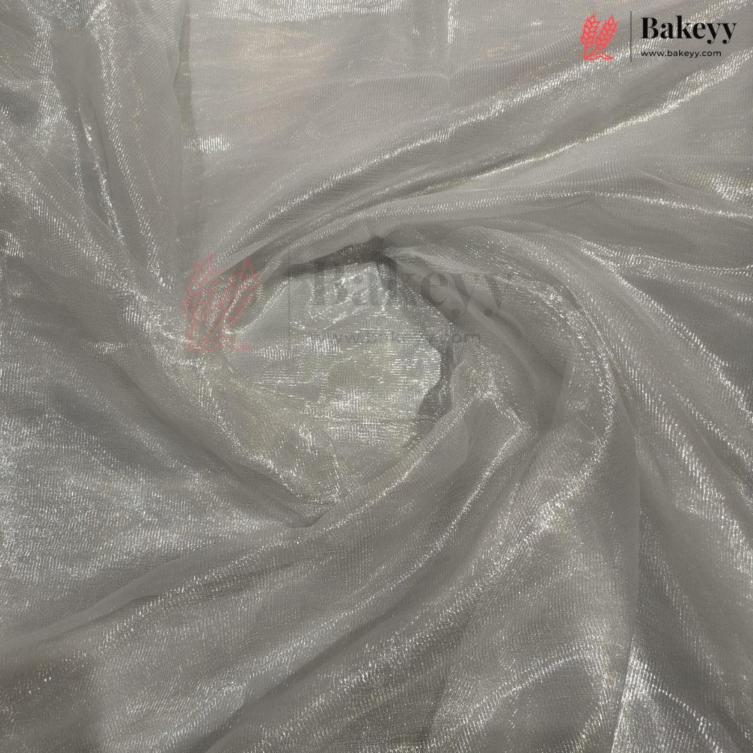 Shimmering silver organza fabric with a metallic reflective finish, draped in a swirl pattern. A Bakeyy brand logo is visible in the top right corner.