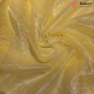 Yellow sheer mesh fabric with a subtle metallic sheen, swirled and draped to show its texture and transparency. A Bakeyy brand logo is visible in the top right corner.